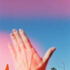 Hand raised in front of a blurred outdoor scene with trees and a clear sky, light leak.