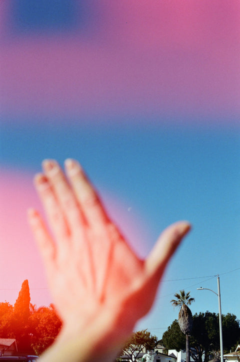 Hand raised in front of a blurred outdoor scene with trees and a clear sky, light leak.